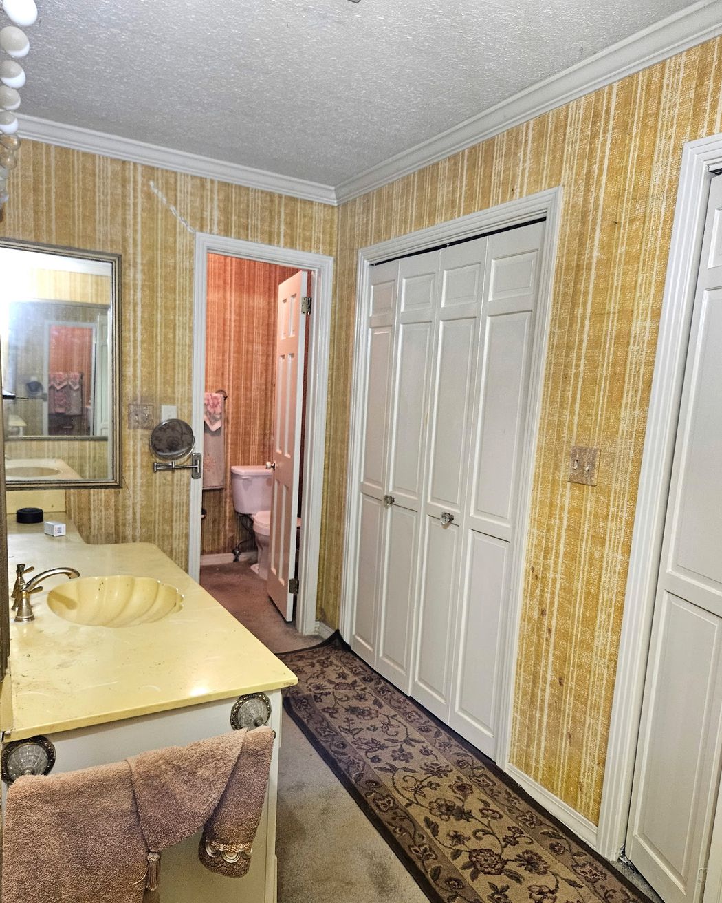 Bath with a cultured-marble vanity and shell sink, striped wallpaper, a run of white closet doors, and a doorway to a separate toilet room