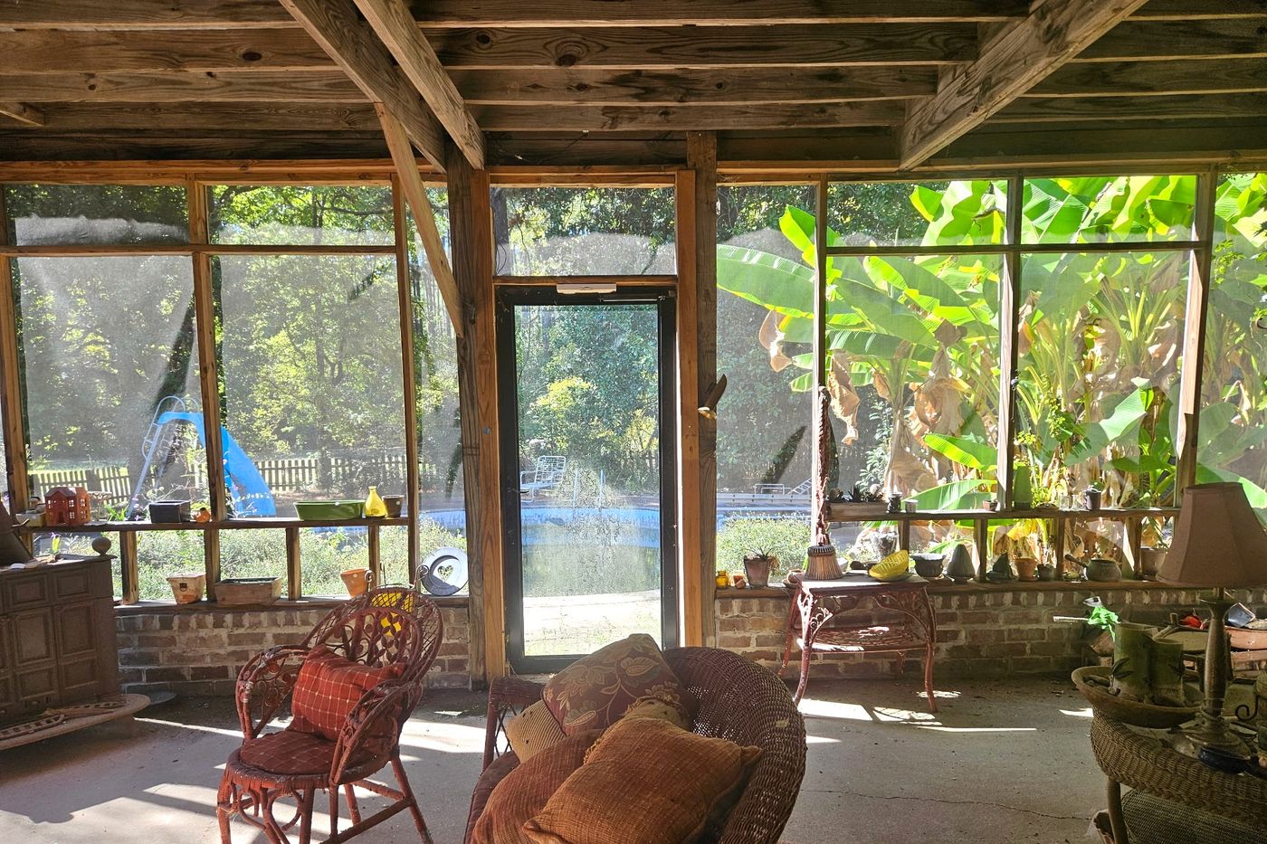 Screened porch with exposed wood ceiling framing, a brick knee wall, and wicker furniture, with the pool and banana plants visible through the screens