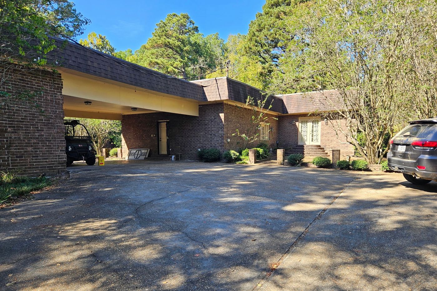 Brick wing of the house with a deep covered carport opening onto a broad paved drive, tall pines behind